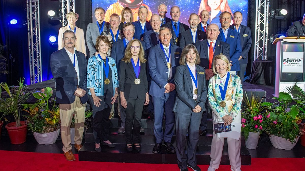 twenty two well dressed people wearing medals stand on a stage surrounded by potted plants