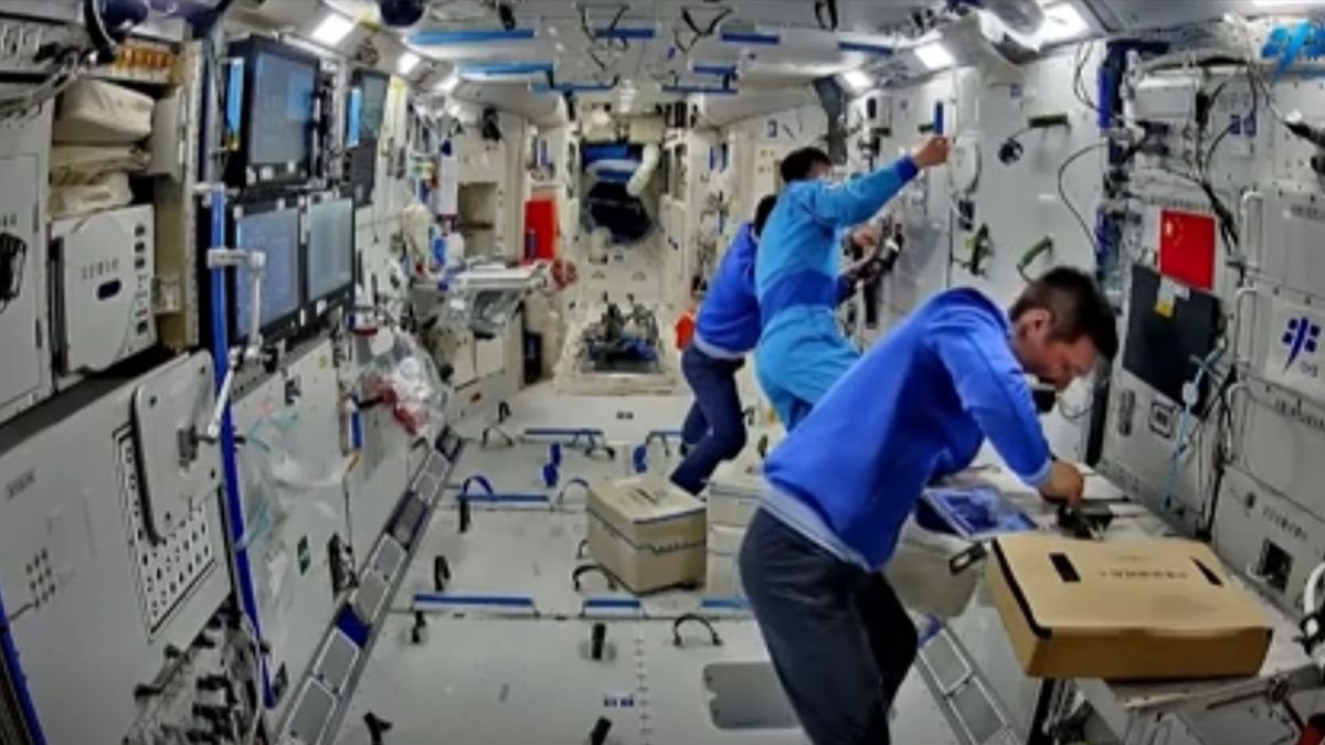three astronauts in blue flight suits work inside a white walled space station