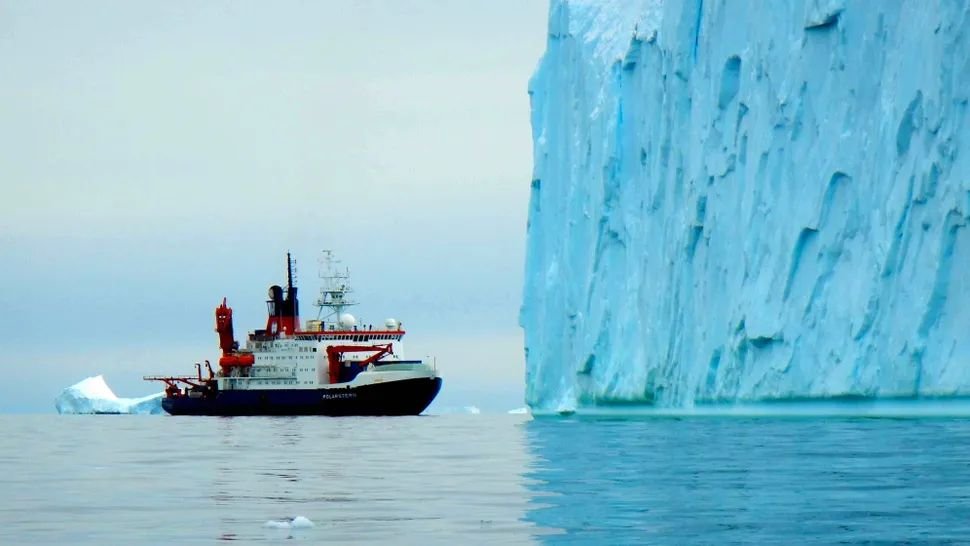 A decent sized boat floats in ocean water next to the vertical incline of an ice cliff