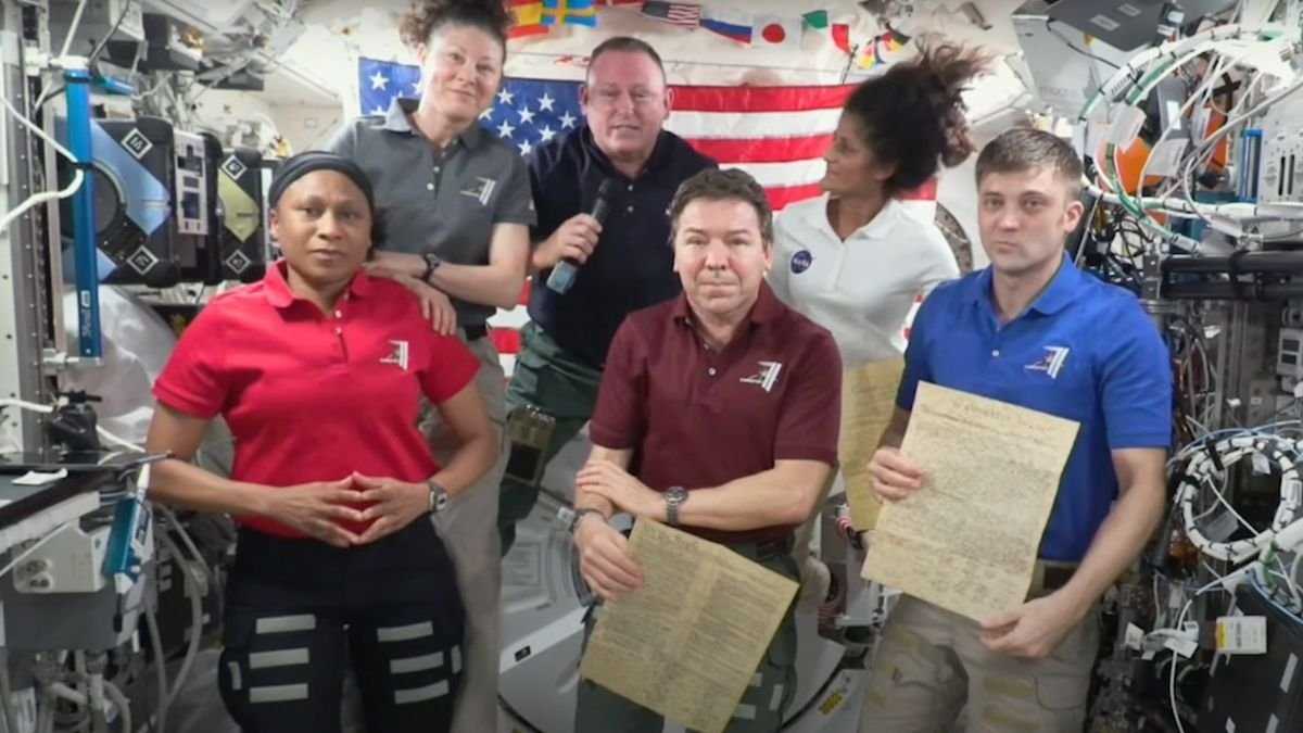 four men and two women float aboard the international space station with an american flag in the background