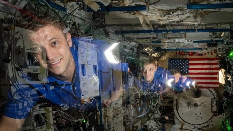 a timelapse image showing an astronaut moving from the back to the front of a module in the international space station at back is an american flag