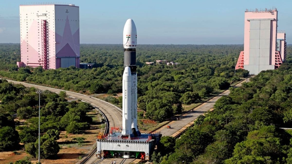 a white rocket with two white side boosters stands on a grey metal platform fixed on a track at the splitting toward two pink decorated hanger buildings