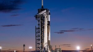 a black and white rocket topped by a white space capsule stand on a launch pad under a darkening evening sky