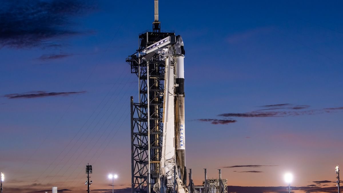 a black and white rocket topped by a white space capsule stand on a launch pad under a darkening evening sky