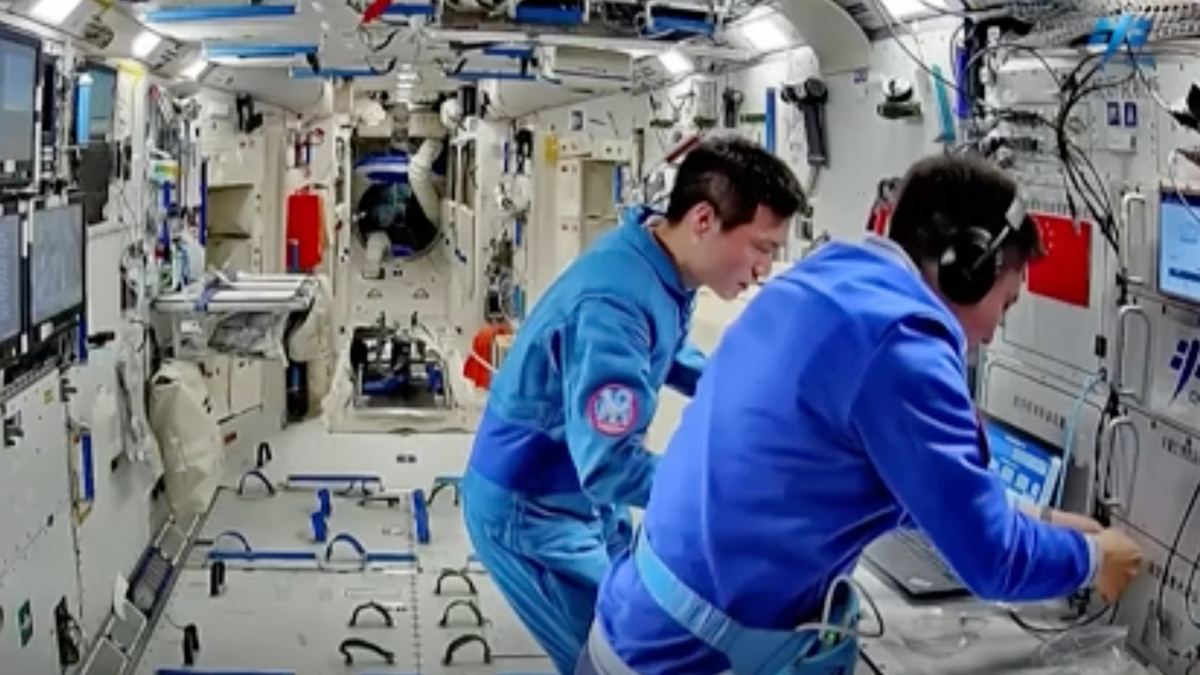 two astronauts in blue flight suits look at a laptop aboard a white-walled space station