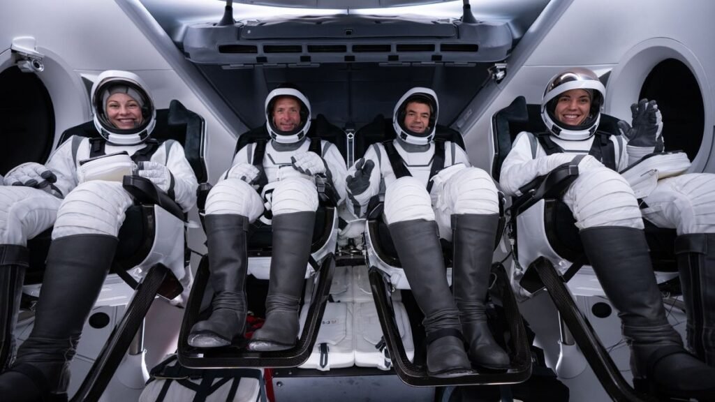 four smiling people in black-and-white spacesuits sit in a space capsule