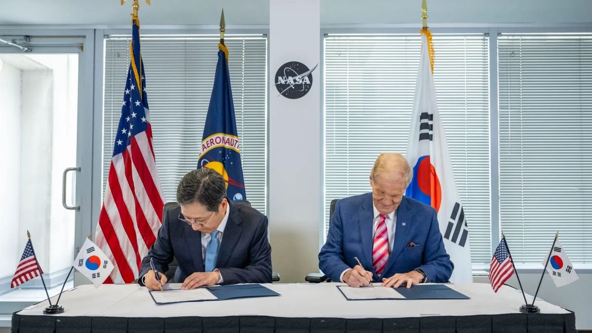 Two men in suits sit at a table in front of some flags and a building support beam sporting a NASA logo. They are both signing their own piece of paper held by a blue folder.