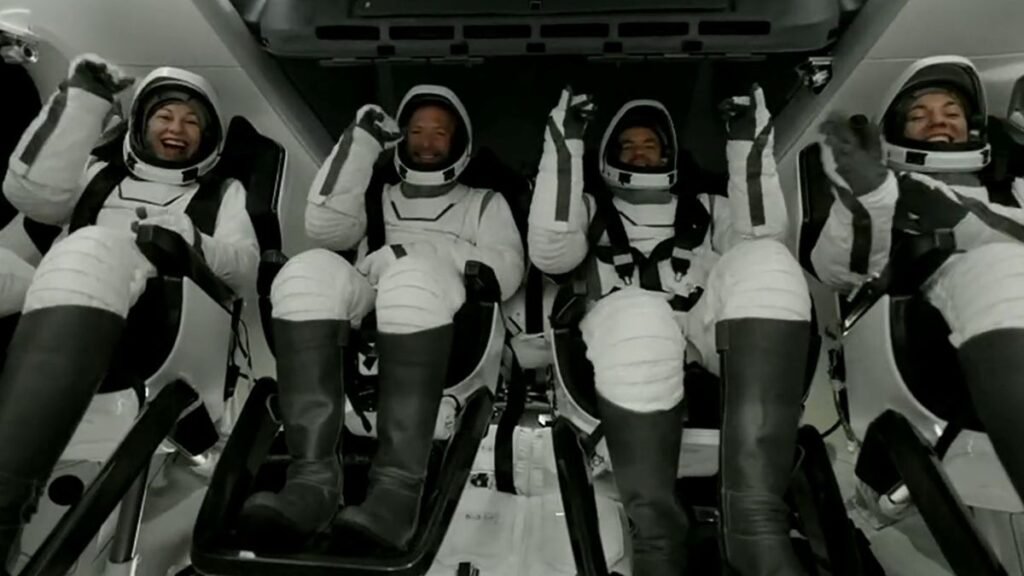 four astronauts in white spacesuits smile and wave inside a cockpit