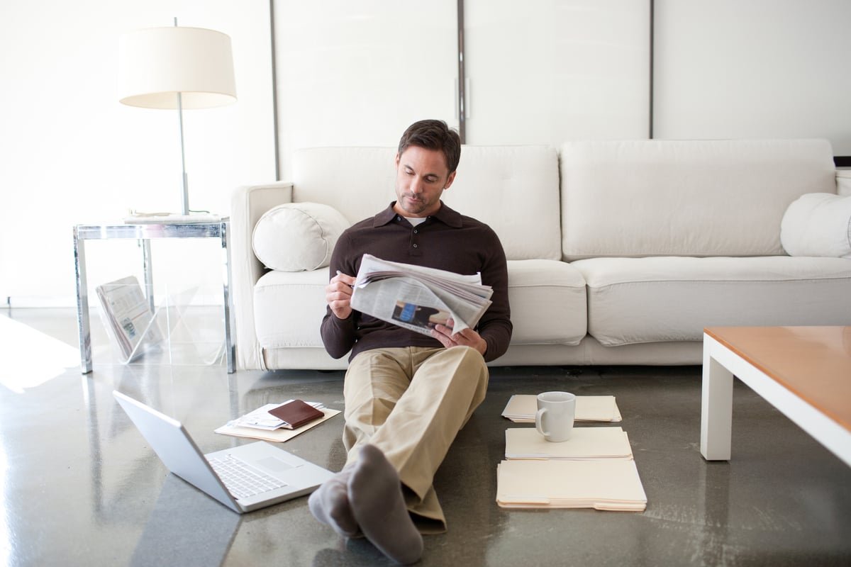 An investor sitting against a couch reading a newspaper, while their laptop and  sets of files are on the floor.