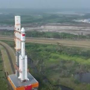 a white rocket rolls out to its launch pad on a track that passes greenery and a small lake