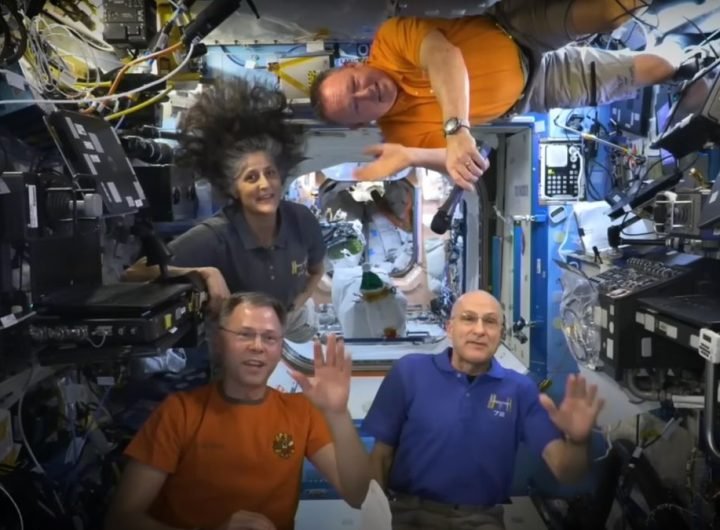 three men and one woman wave from inside the international space station