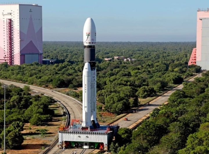 a white rocket with two white side boosters stands on a grey metal platform fixed on a track at the splitting toward two pink decorated hanger buildings