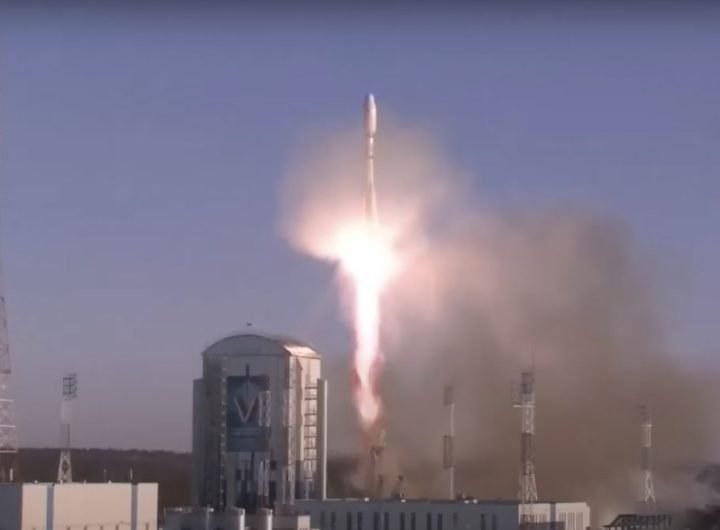 A rocket blasts off near launch facilities and tall lightning towers.