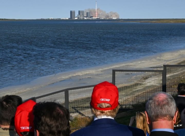 a crowd of people, including a man in a red baseball cap, watch a rocket launch from across a stretch of water