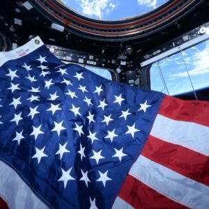 an american flag floats in a small circular room lined with windows. through the windows, earth can be seen far below