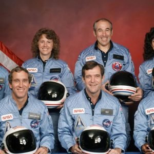 seven astronauts in blue flight suits sit for a portrait while holding their helmets