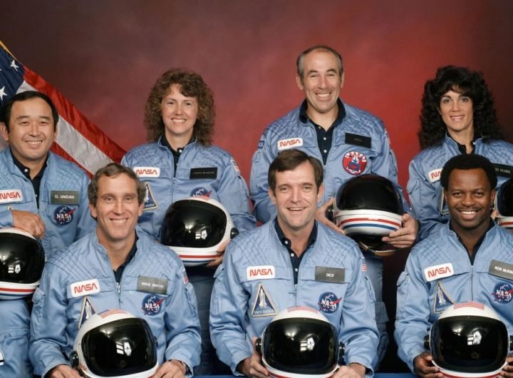 seven astronauts in blue flight suits sit for a portrait while holding their helmets